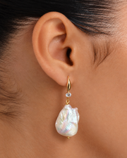 A close-up of a large baroque pearl earring with an iridescent sheen, suspended from a gold-tone hook.